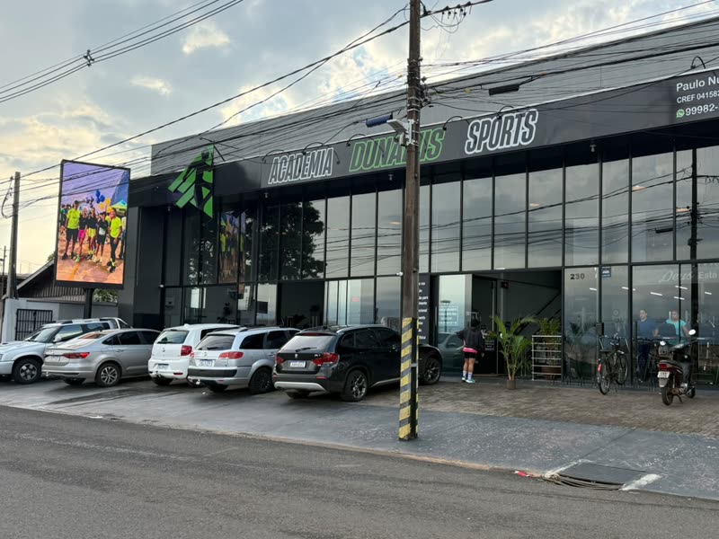 Painel de LED vertical da Soligado Mídia, aceso, instalado num poste na esquina da fachada da Academia Dunamis Sports, com carros estacionados e a avenida em primeiro plano, em Paiçandu