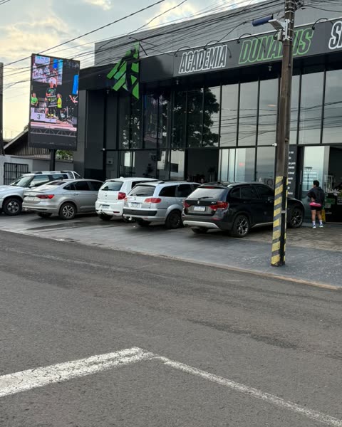 Vista da rua em frente ao painel: o painel de LED aceso à esquerda, carros estacionados na fachada da academia e a faixa de pedestres em primeiro plano